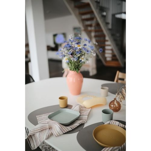 Een gedekte tafel met mushie voedingsbenodigdheden voor baby's en peuters, waaronder bekers, borden en een slab, met een vaas bloemen.