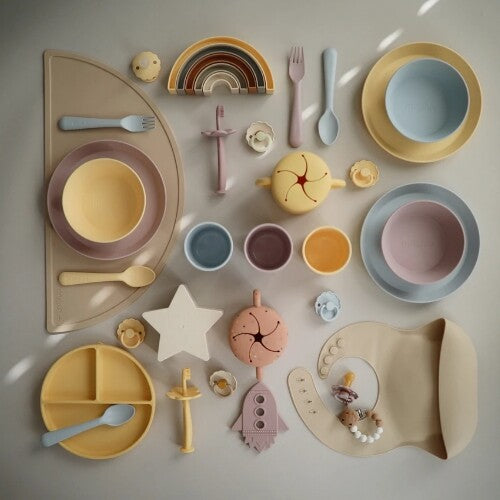 Een bovenaanzicht van een uitgebreide collectie mushie baby- en peuterproducten, waaronder servies, slabbetjes en speelgoed.
