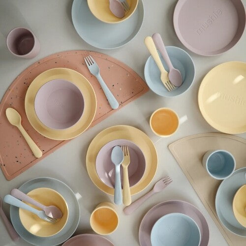 Een bovenaanzicht van diverse mushie baby- en peuterservies onderdelen in pastelkleuren, deels op placemats.