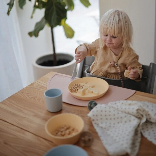 Een peuter eet met een vork van een geel kinderbord in een kinderstoel aan tafel.