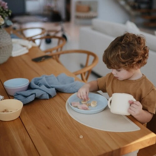Een jong kind eet van een mushie kinderbord aan een houten tafel.