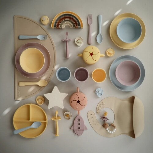 Een uitgebreide collectie mushie babyproducten in pastelkleuren, waaronder voedingsgerei, speelgoed en fopspenen.