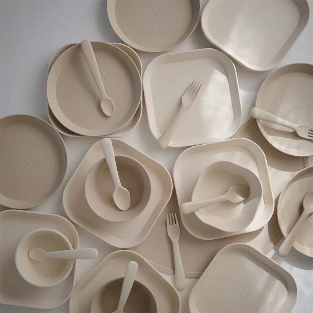 Een bovenaanzicht van een collectie beige mushie kinderborden, kommen en bestek.