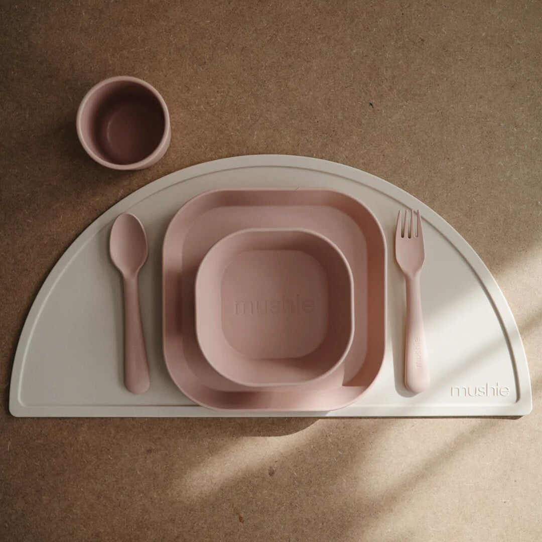 Een mushie babyvoedingsset in blush roze met een vierkant bord, kom, beker, lepel en vork op een lichtgrijze placemat.