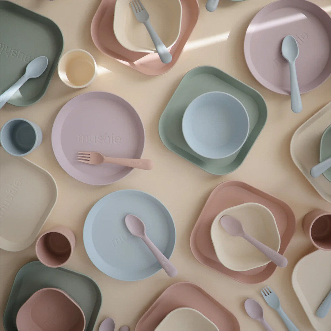 Een overzicht van diverse mushie babyvoedingsartikelen in pastelkleuren, waaronder borden, kommen, bekers en bestek.