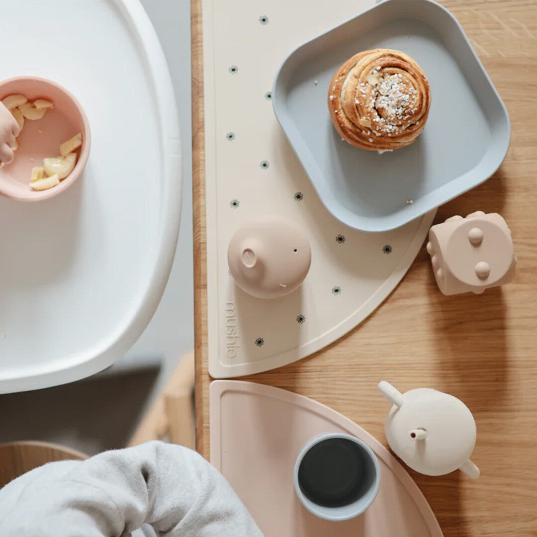 Een tafelsetting met een lichtgrijs mushie kinderbord met een kaneelbroodje en andere babyvoeding benodigdheden.
