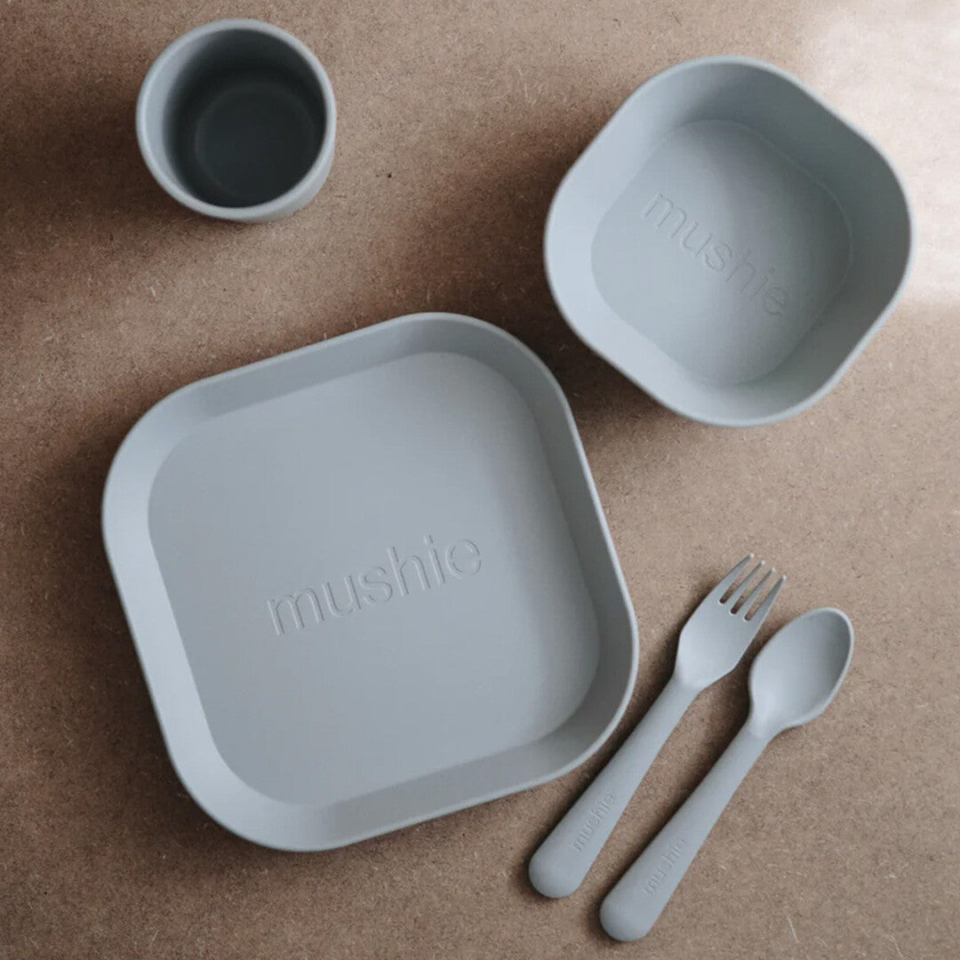 Een complete lichtgrijze mushie babyvoeding set met een bord, kom, beker en bestek.