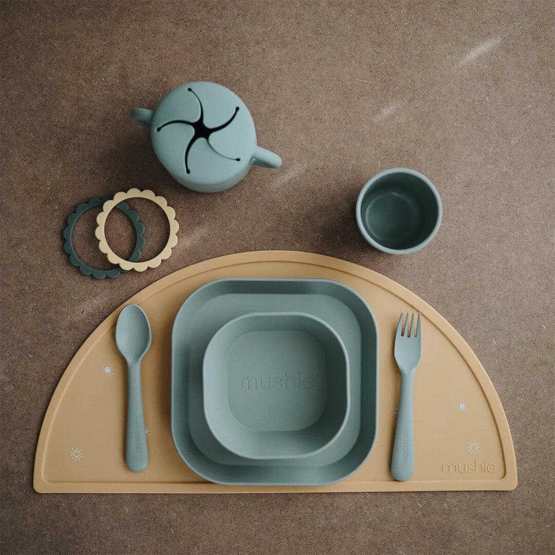 Een set mushie babyvoedingsbenodigdheden in groen en beige, inclusief bord, kom, beker en bestek, op een placemat.