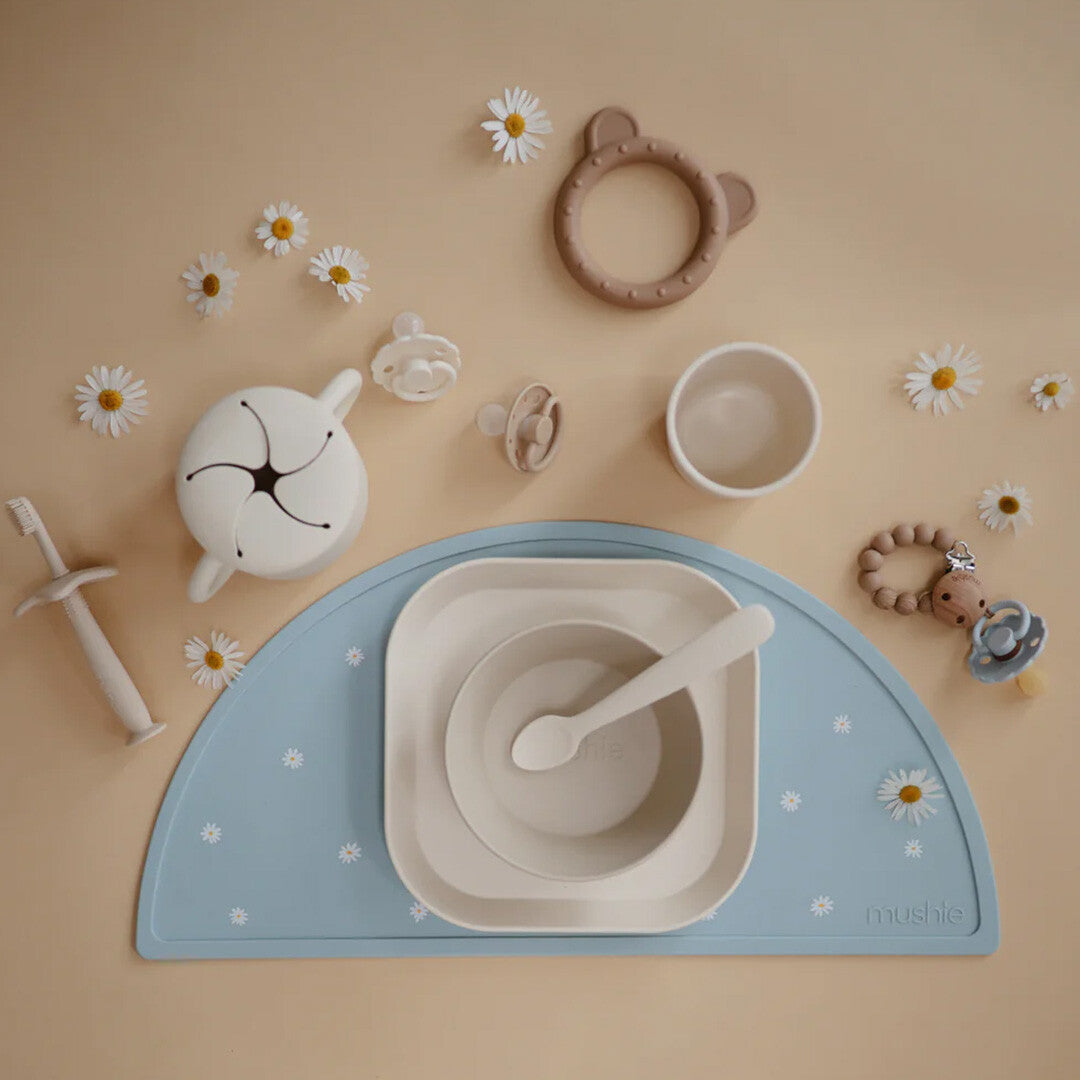 Een flat lay van mushie babyvoedingsbenodigdheden, waaronder borden, op een blauwe placemat met madeliefjes.