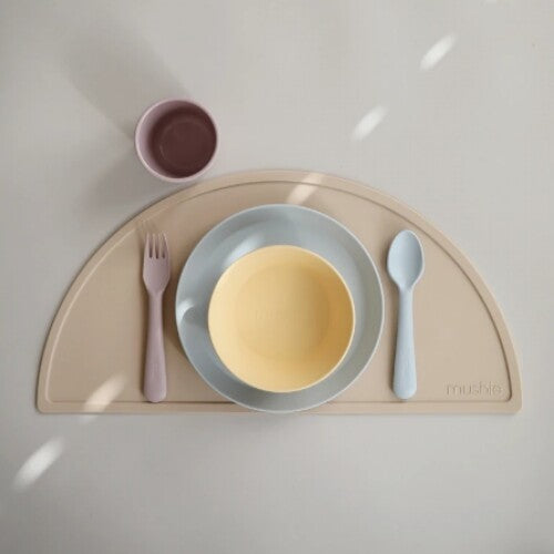 Een mushie kinderservies set met een gele kom, blauwe borden en bestek op een beige placemat.