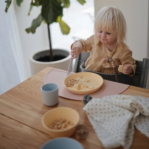 Een peuter eet uit een gele kinderkom op een roze placemat aan een houten tafel.