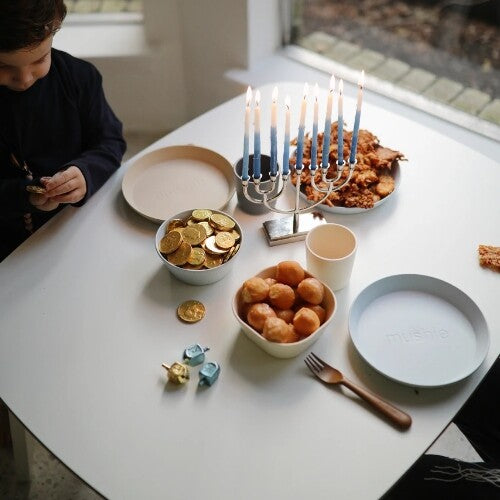 Een kind aan tafel met mushie kinderservies, eten en een brandende menorah.