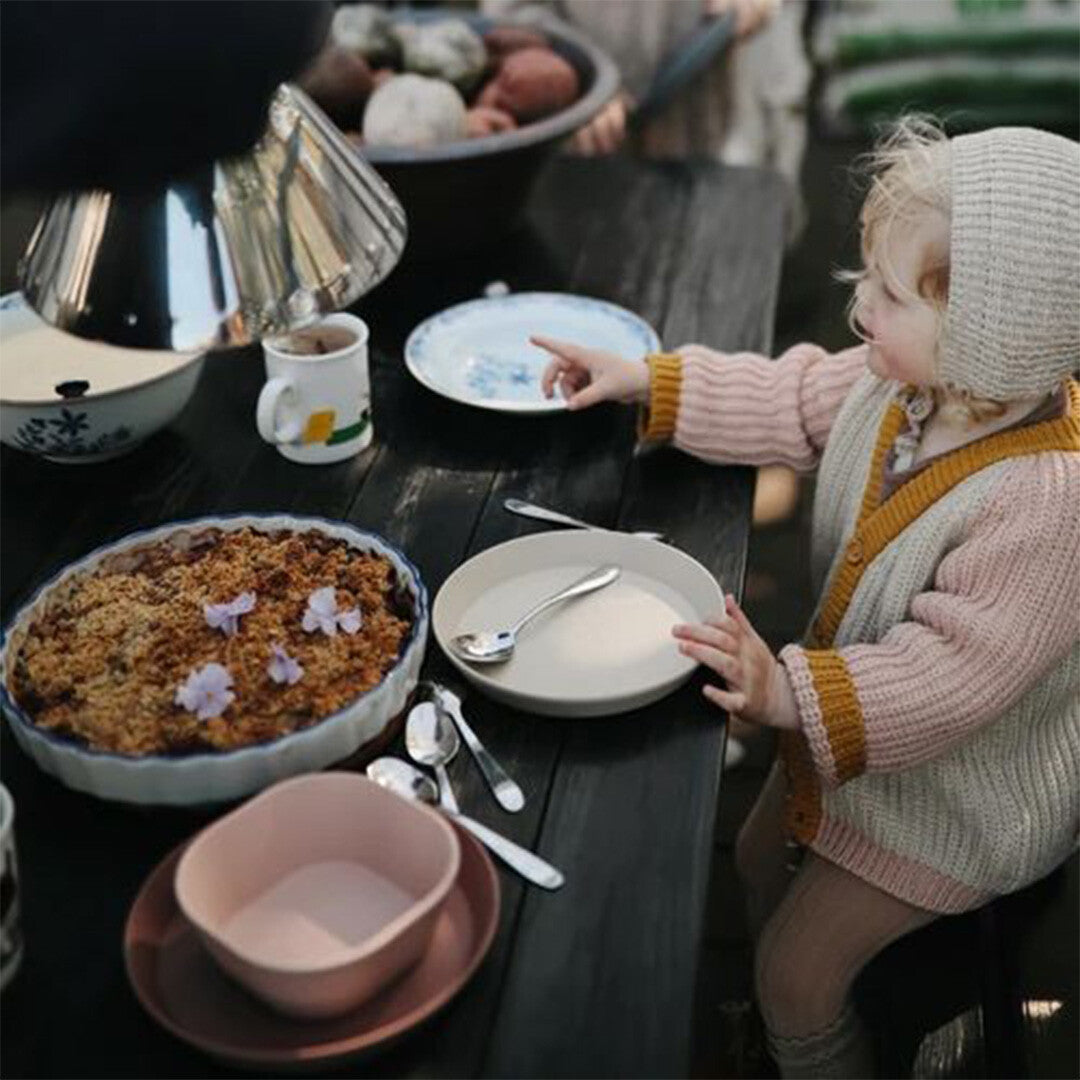 Een kind zit aan tafel met een roze kinderkom en andere servieswaren.