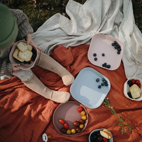 Een kind eet buiten van een mushie kinderkom, met meerdere gekleurde mushie kommen en borden met fruit en snacks op een picknickkleed.