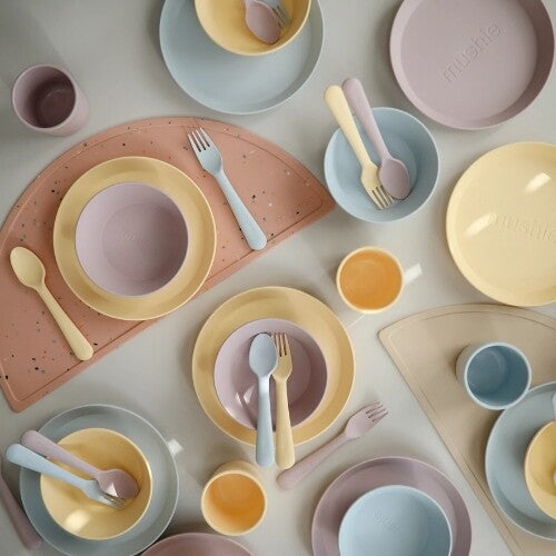 Een schuin bovenaanzicht van mushie kinderborden, kommen, bekers, bestek en placemats in pastelkleuren.