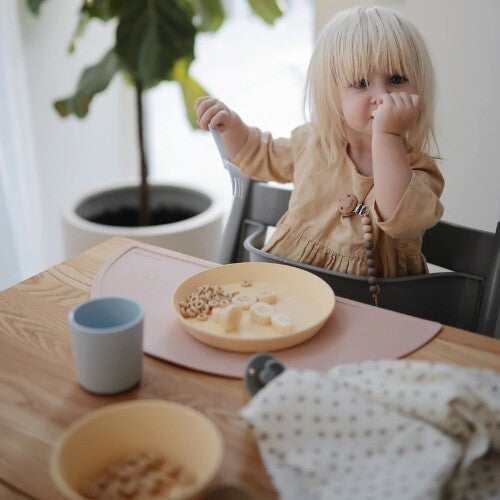 Een kind zit in een kinderstoel aan tafel met een geel bord met eten en een blauwe beker op een roze placemat.
