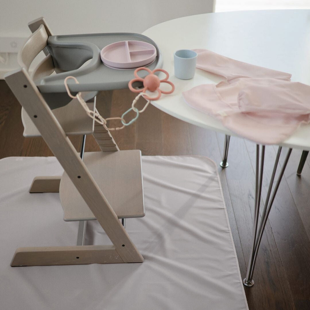 Beige mushie knoeimat onder een kinderstoel met babyvoedingsaccessoires op tafel.