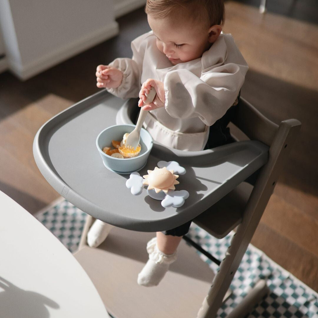 Baby in een kinderstoel die eet, met de mushie knoeimat met geruit patroon op de vloer.