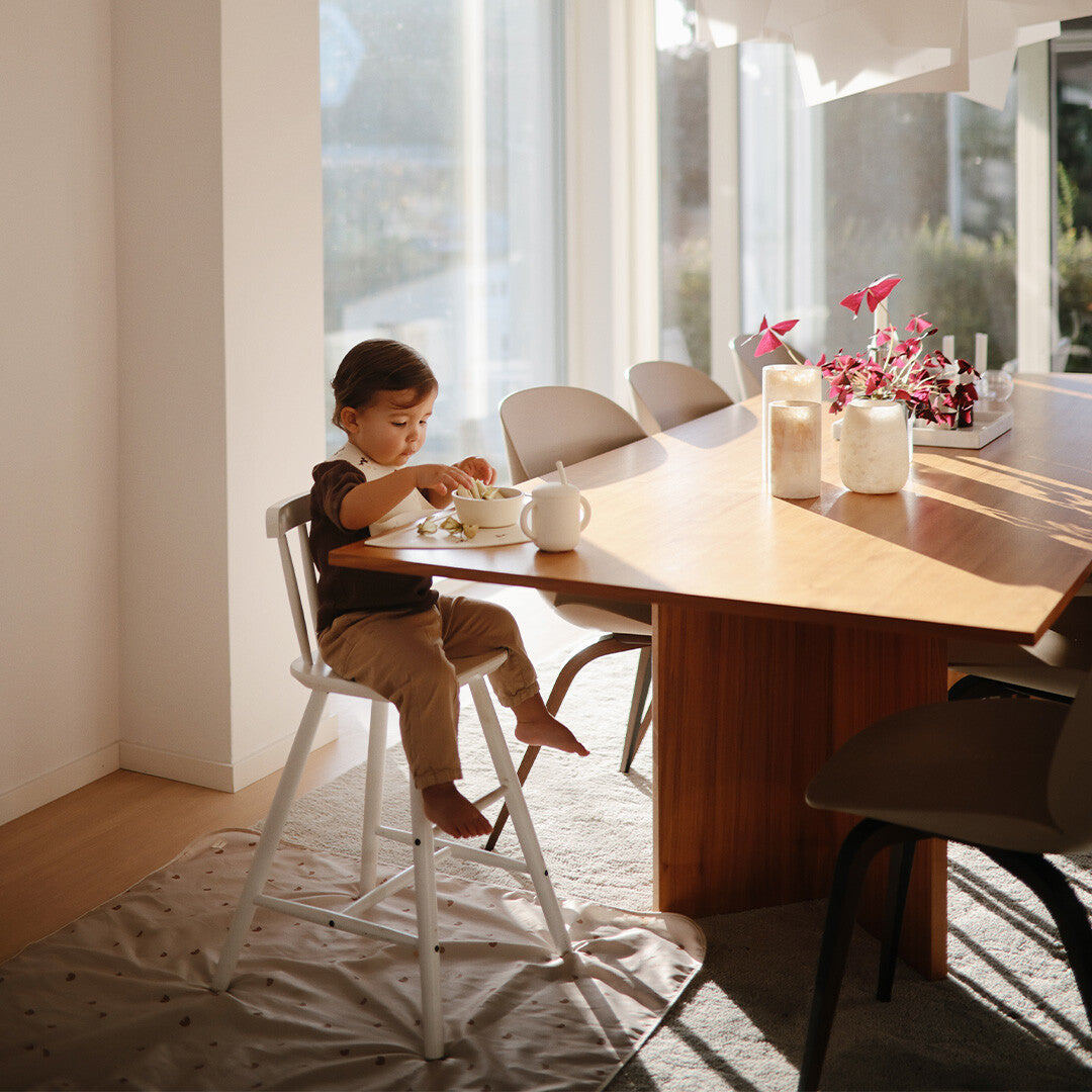 Peuter eet aan tafel in een kinderstoel, met de mushie knoeimat op de vloer.