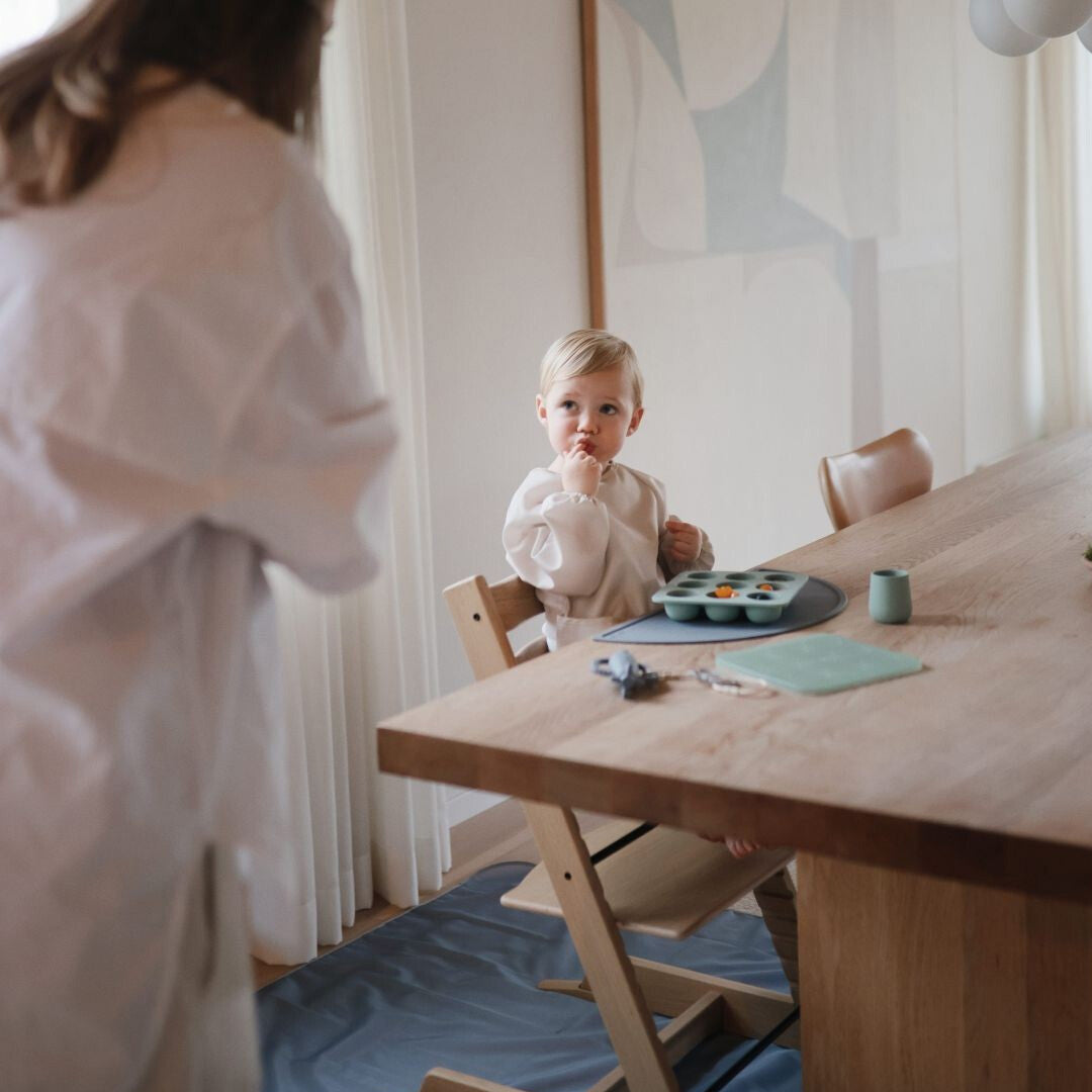 Een baby zit in een kinderstoel aan tafel, met een blauwe mushie knoeimat op de vloer eronder.
