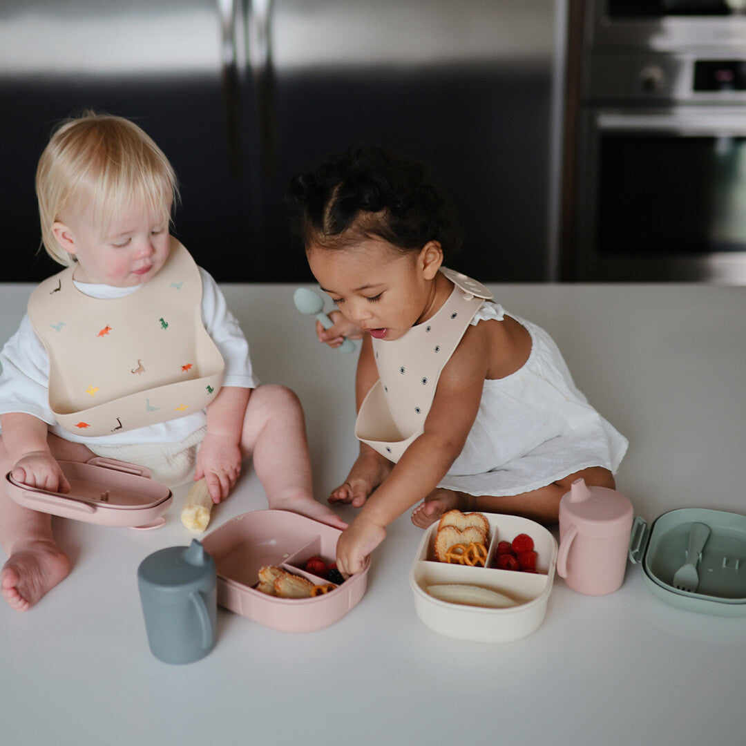 Twee baby's met slabbetjes eten uit Mushie lunchboxen en drinken uit bekers op een aanrecht.