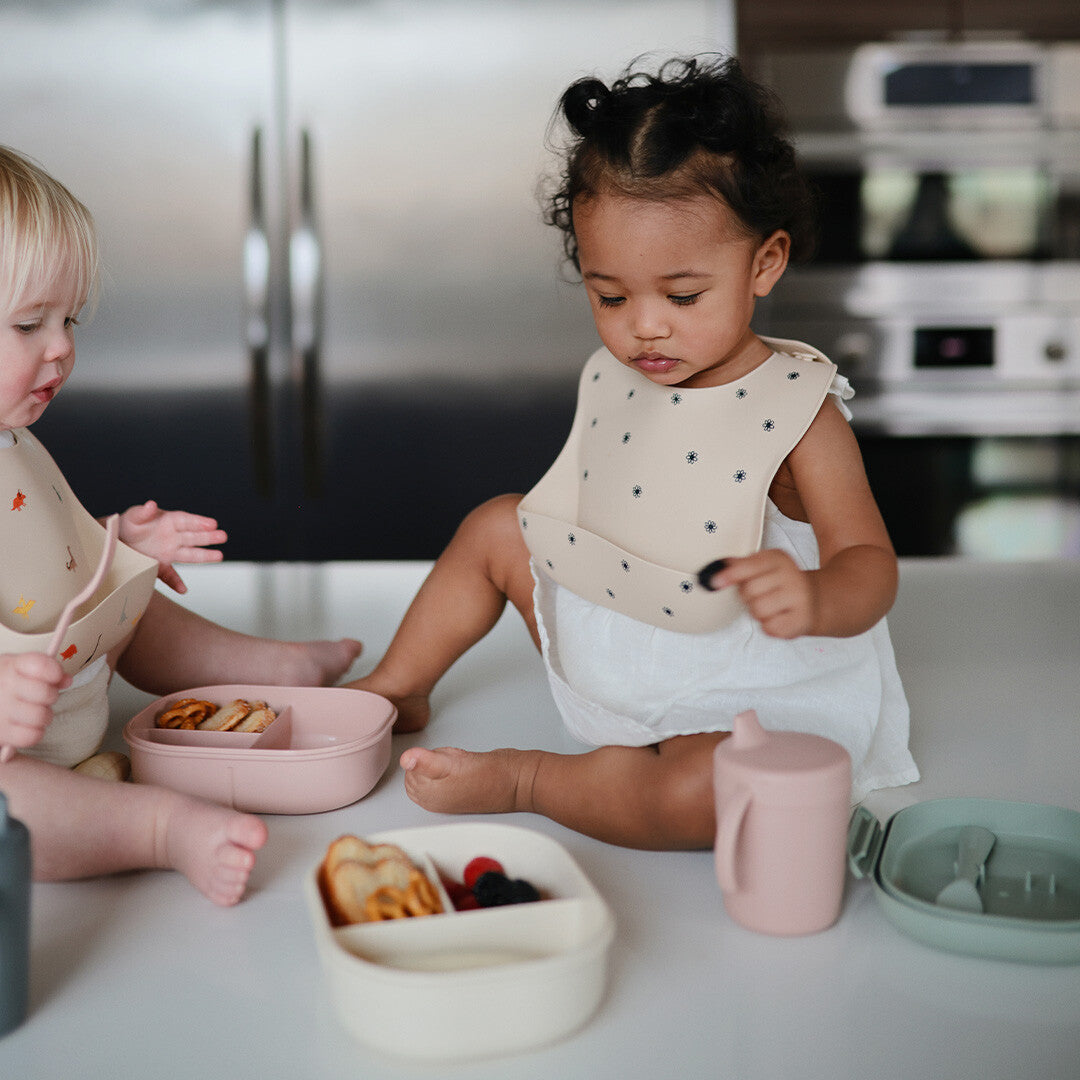Twee baby's met mushie slabbetjes en lunchboxen op een aanrecht, waarbij één kind in een lunchbox kijkt.