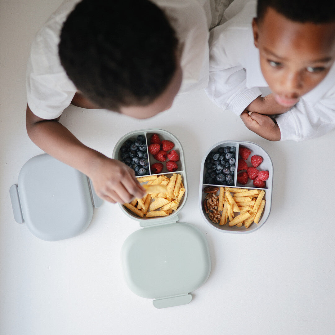 Twee kinderen kijken naar open mushie lunchboxen gevuld met fruit en snacks.