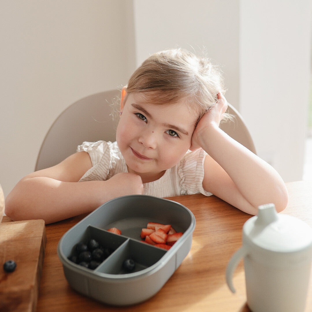 Een meisje zit aan tafel met de open Mushie grijze lunchbox gevuld met fruit.