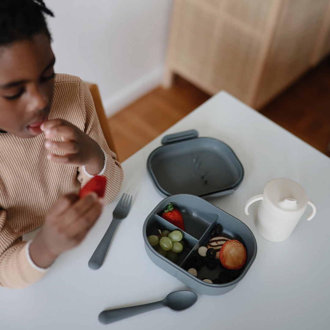 Een kind eet aan tafel met de open Mushie grijze lunchbox gevuld met diverse snacks en fruit.