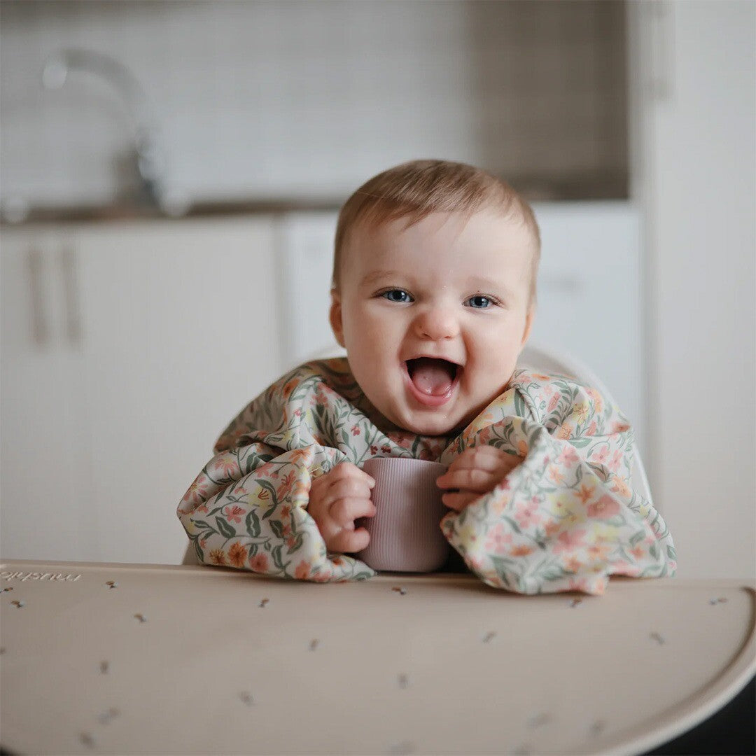 Een lachende baby in een kinderstoel draagt de mushie mouwslab met bloemenpatroon en houdt een beker vast.