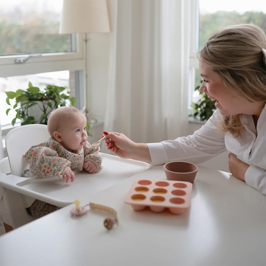 Een baby in een kinderstoel met de mushie mouwslab wordt gevoed door een volwassene aan tafel.