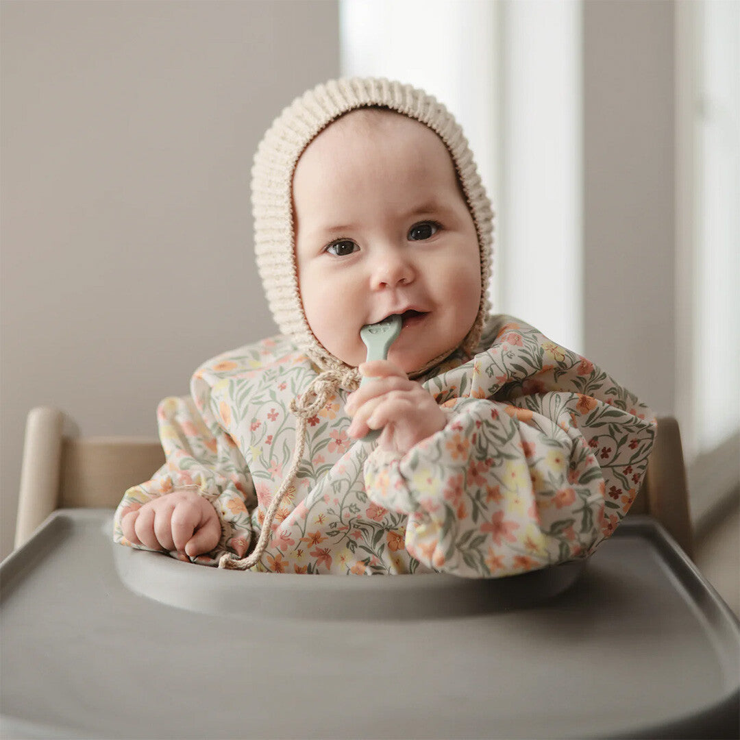 Een baby met een mutsje draagt de mushie mouwslab met bloemenpatroon en houdt een lepel in de mond.