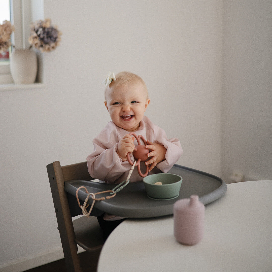 Een lachende baby draagt een Mushie roze mouwslab met regenboogpatroon in een kinderstoel.