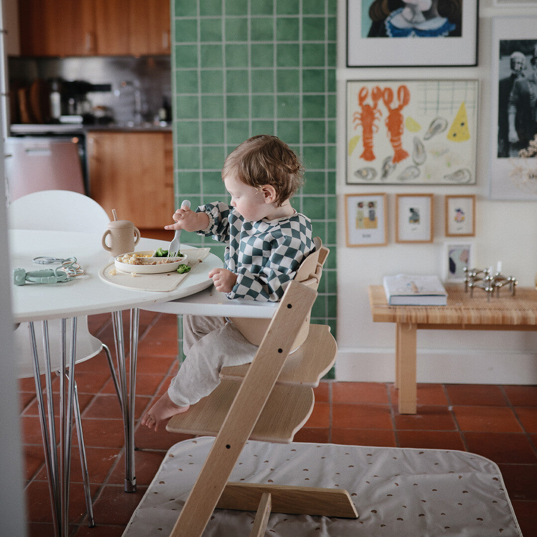 Een baby draagt een geruite mouwslab en eet met een vork aan een tafel in een kinderstoel.