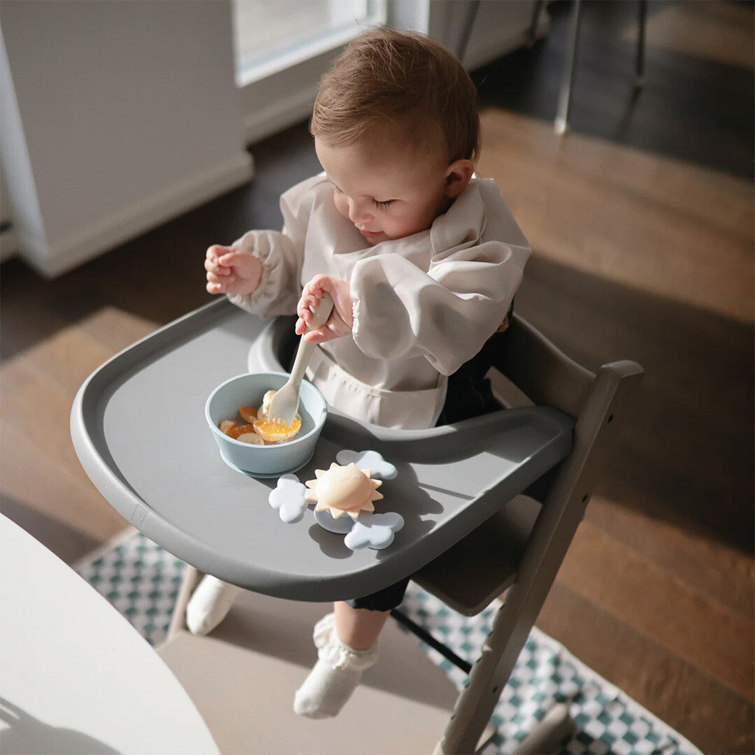 Een baby draagt een lichtgekleurde mouwslab en eet uit een kom in een kinderstoel.