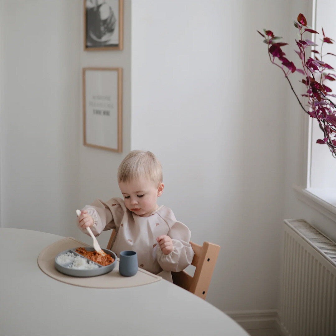 Een baby met een lichtgekleurde mouwslab met regenboogpatroon eet aan tafel in een kinderstoel.