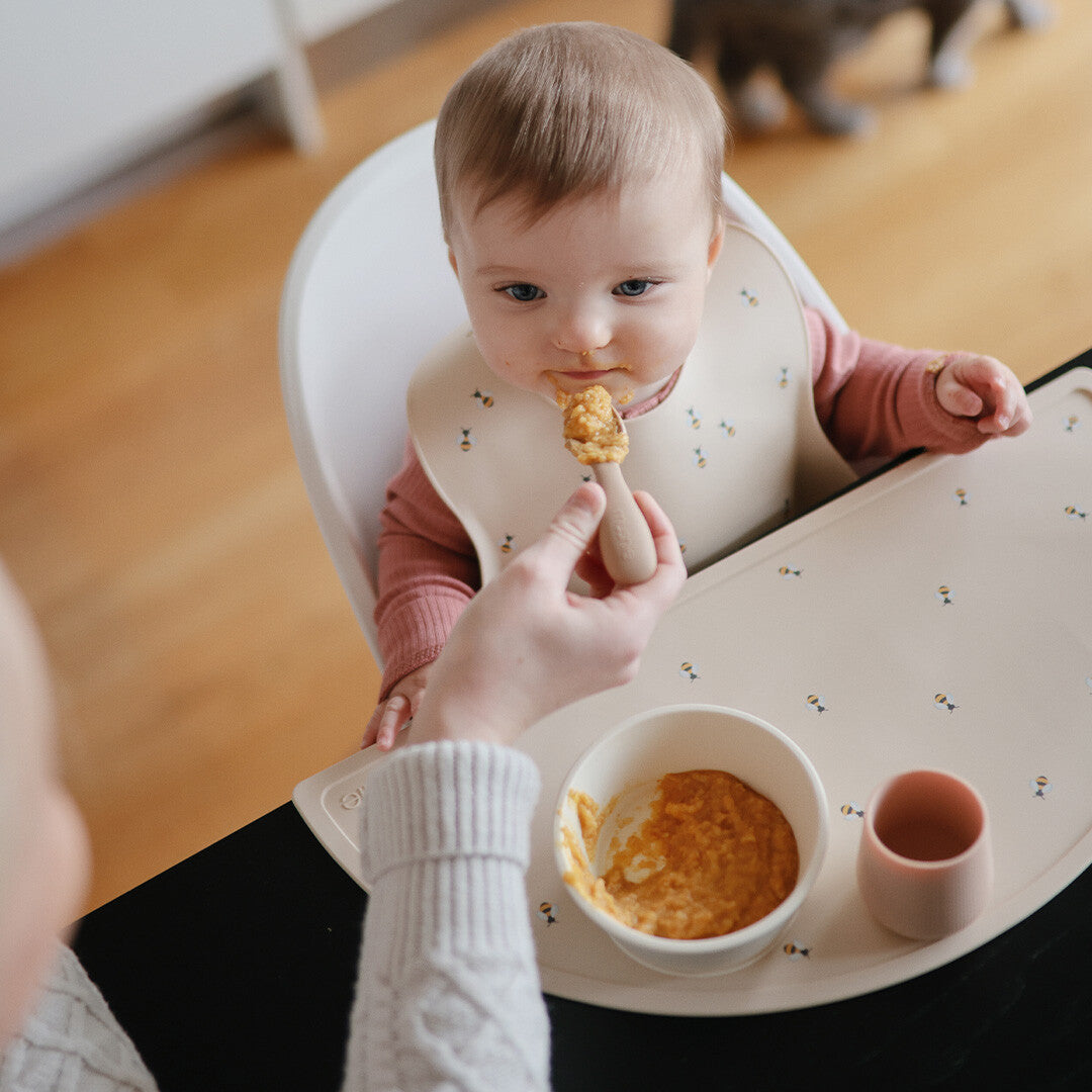 Een baby in een kinderstoel wordt gevoed, met een mushie blush peuterbeker op de placemat.