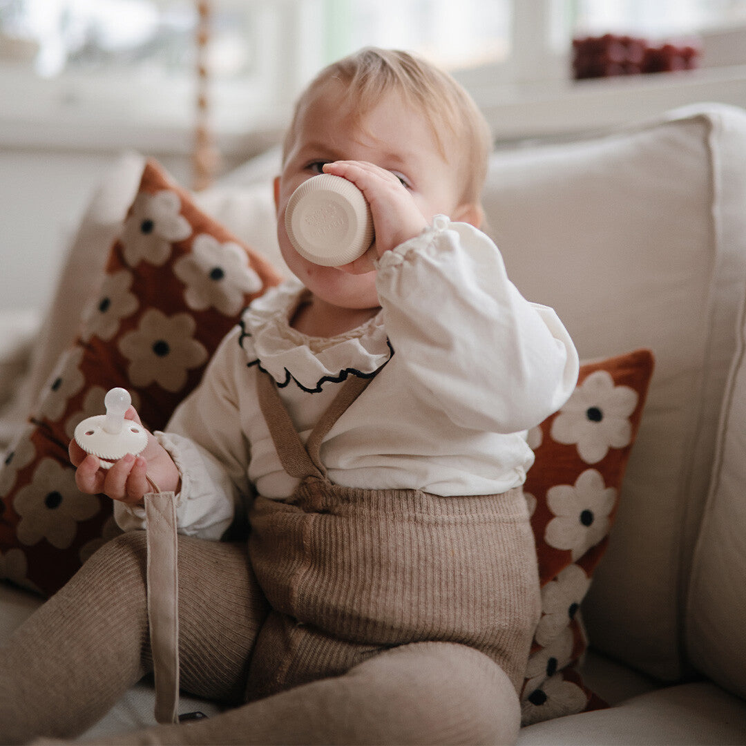Een baby drinkt uit een mushie peuterbeker in de kleur shifting sand, zittend op een bank.