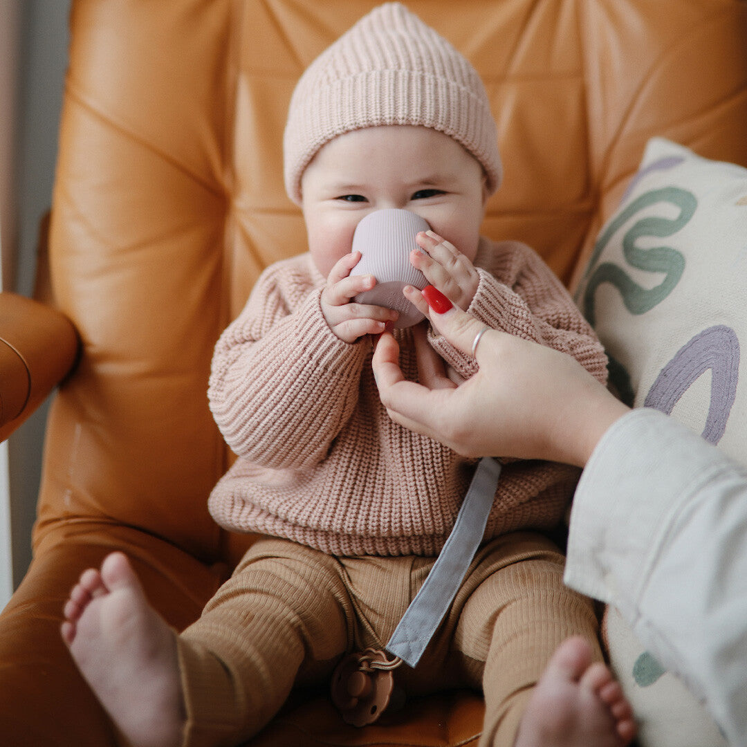 Een baby met een roze muts en trui houdt een mushie peuterbeker in zachtroze siliconen tegen het gezicht.