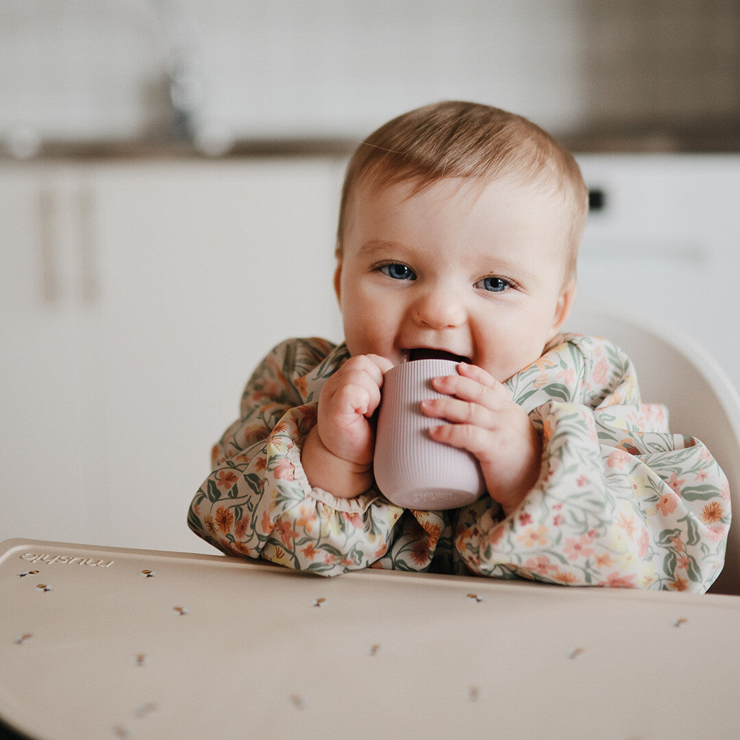 Een lachende baby in een kinderstoel houdt een mushie peuterbeker in zachtroze siliconen vast.