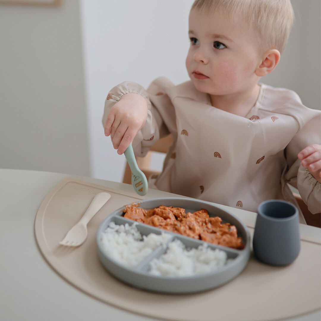 Een peuter in een kinderstoel met een mushie peuterbeker en een bord met eten.