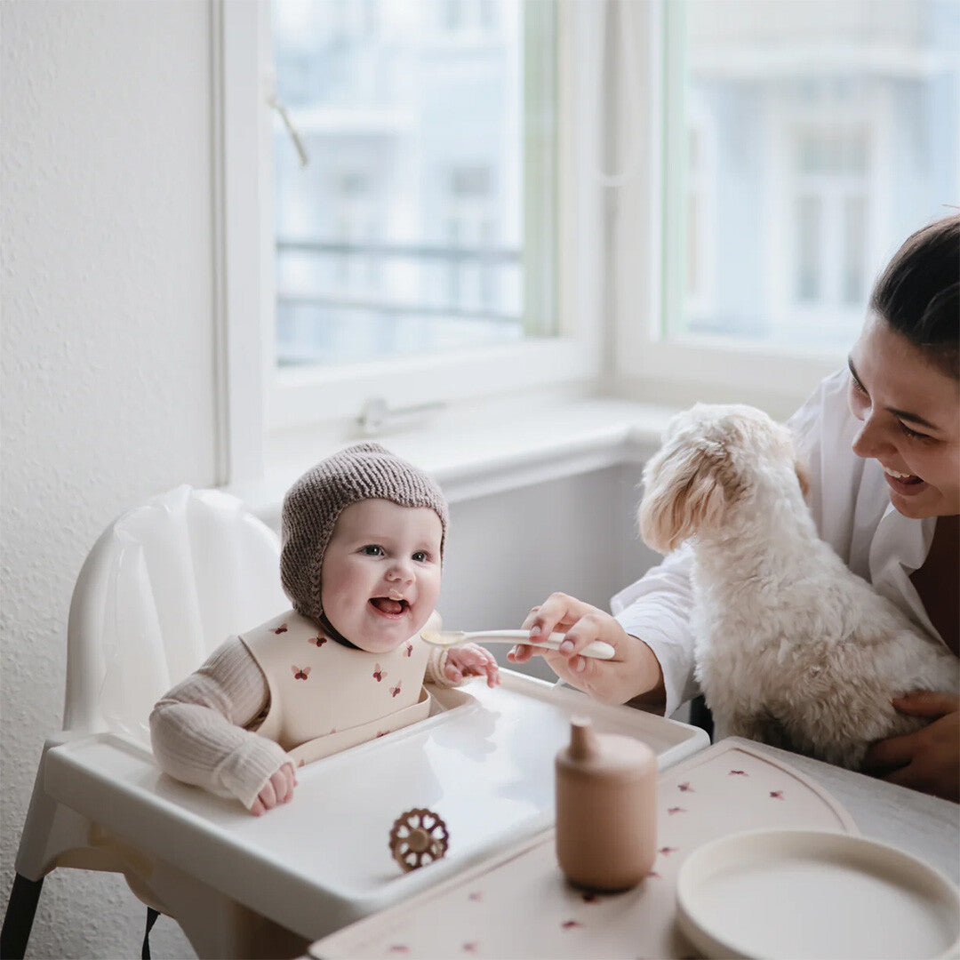 Een baby in een kinderstoel wordt gevoed, met een mushie placemat en slab met vlinderpatroon op de tafel.