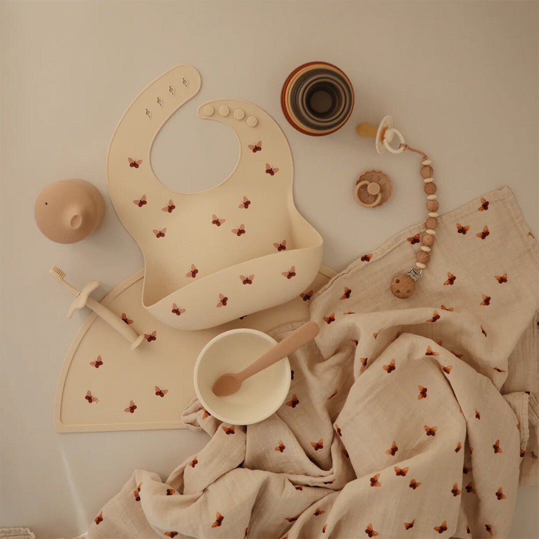 Een collectie mushie babyvoedingsbenodigdheden en accessoires, waaronder een placemat, slab en doek met vlinderpatroon.