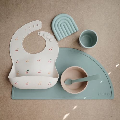 Mushie babyvoedingsset met een kersjes slab, een regenboog bijtspeeltje, een kom, lepel en beker op een Cambridge blue placemat.