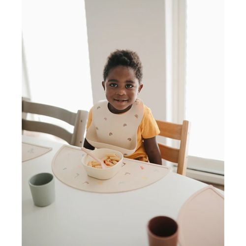 Een lachend kind met een Mushie regenboog slab zit aan tafel met een Mushie placemat en een kommetje eten.