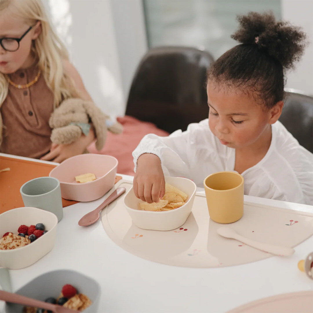 Kind eet snacks aan tafel met een beige placemat met kersenpatroon.