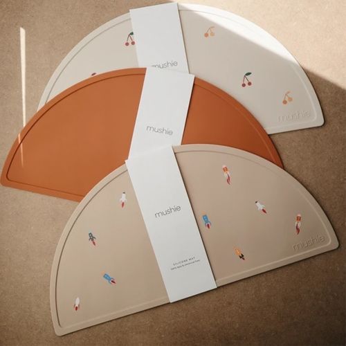 Drie Mushie siliconen placemats in verschillende kleuren en patronen, met labels.