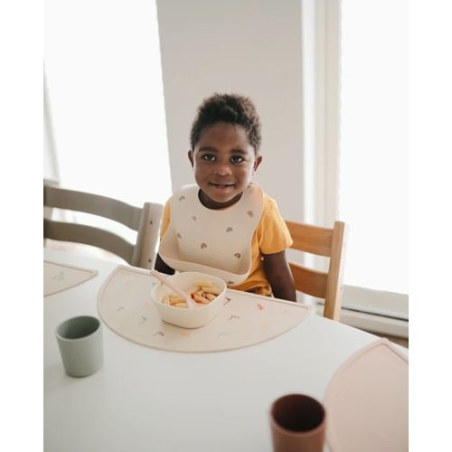 Een lachend kind zit aan tafel met een beige placemat en een kommetje eten.