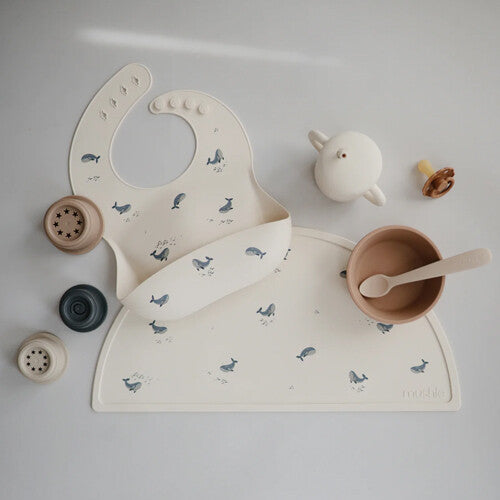Mushie beige siliconen placemat met walvissen, een bijpassend slabbetje en diverse babyvoedingsaccessoires.