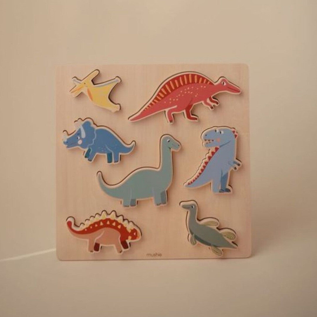 Mushie houten puzzel met zeven kleurrijke dinosaurusvormige stukken op een lichte achtergrond.